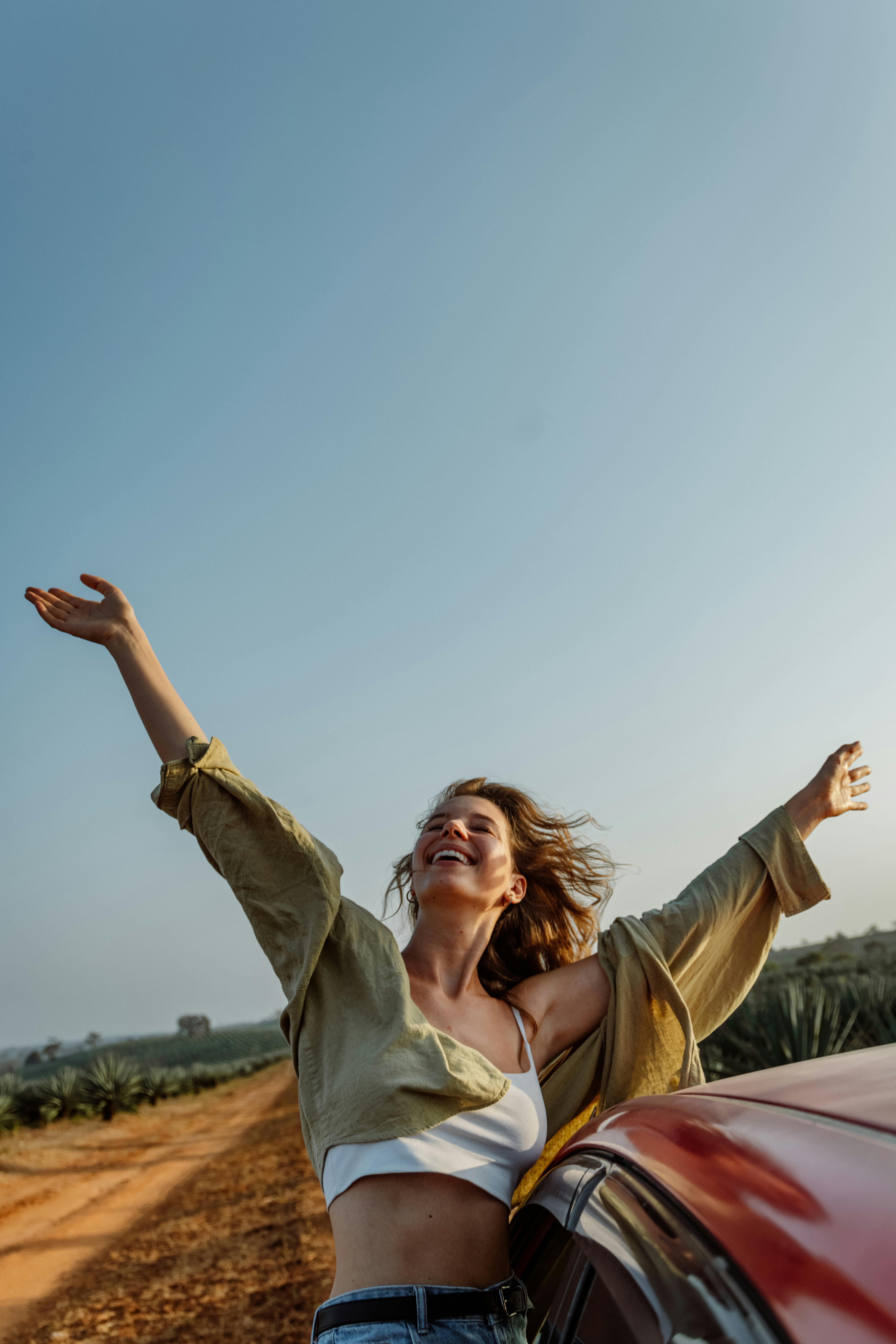 Woman joyfully stretches arms beside car on a sunny rural road, embracing freedom.